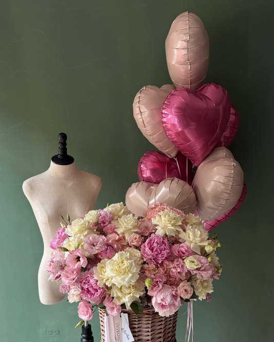 Bouquet of flowers in a basket with heart-shaped balloons against a green wall.
