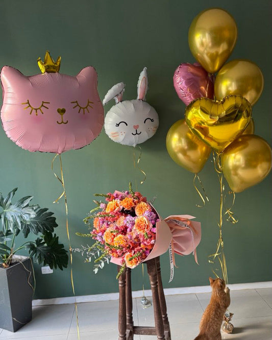 Decorative balloons, flowers, and a cat on a stool against a green wall.