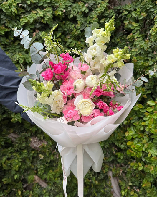 Ramo de flores de estación rosadas y blancas con ranúnculos, claveles, rosas spray y conejitos, envuelto en papel blanco.