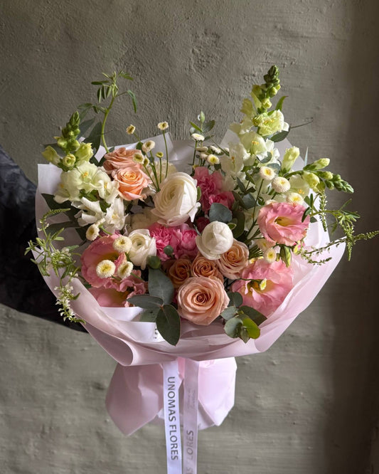 Ramo de ranúnculos, rosas spray y lisianthus en tonos rosados, durazno y crema, envuelto en papel rosa.