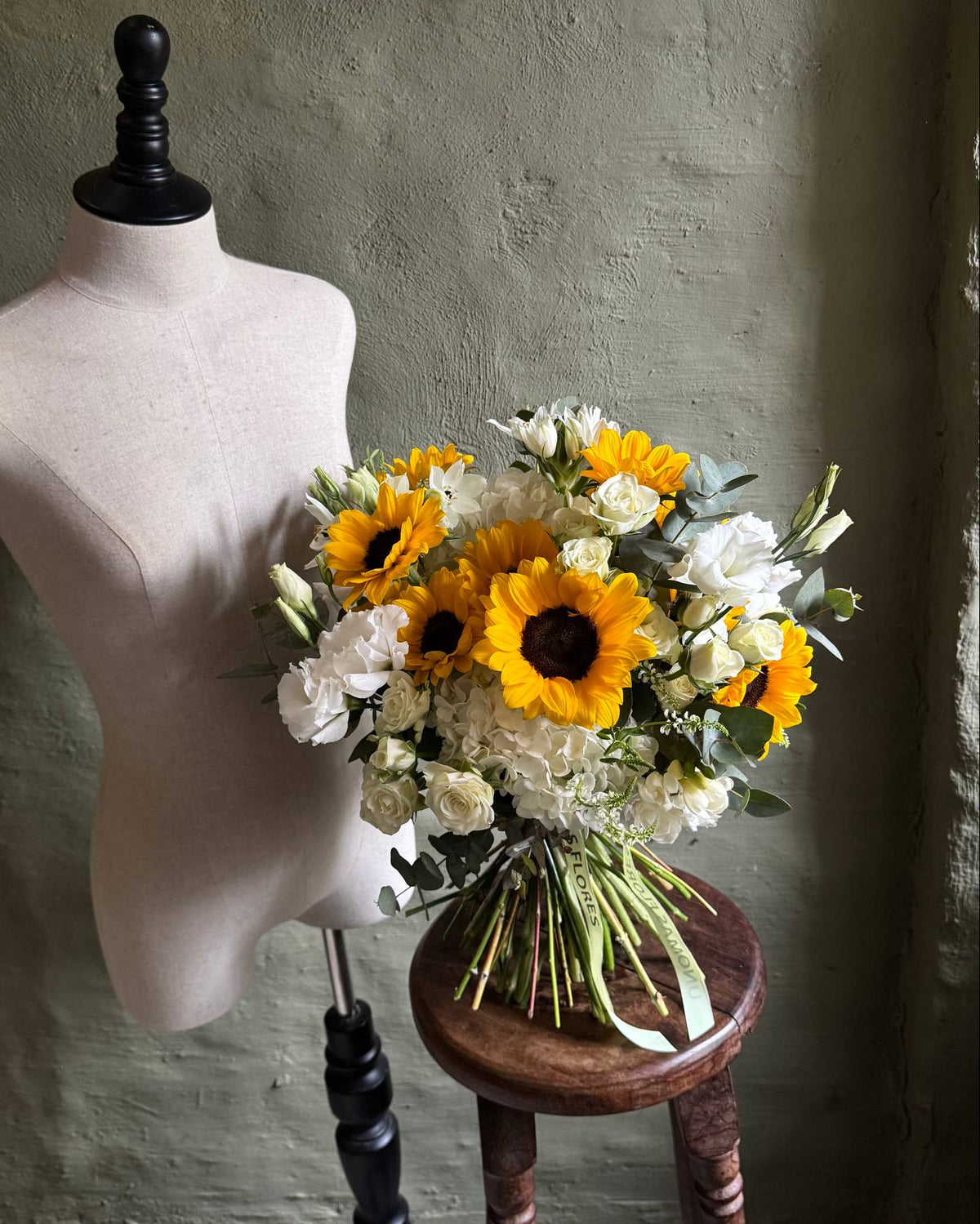 Ramo de girasoles amarillos, hortensias y lisianthus blancos con follaje, atado con una cinta sobre un banquito.