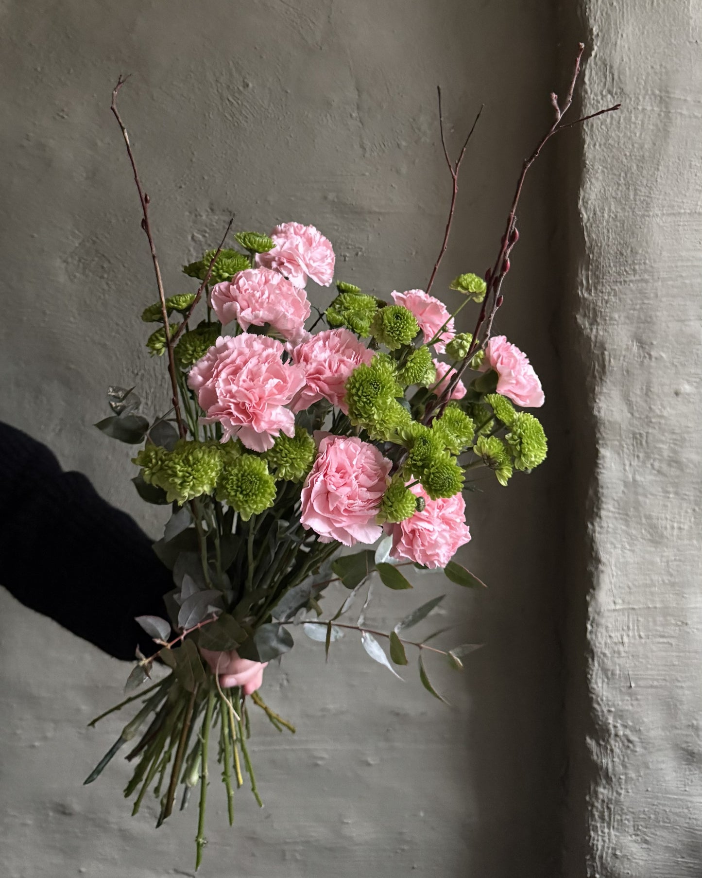 A bouquet of pink carnations and green pom-pom chrysanthemums with eucalyptus stems, held by a hand against a textured gray wall.