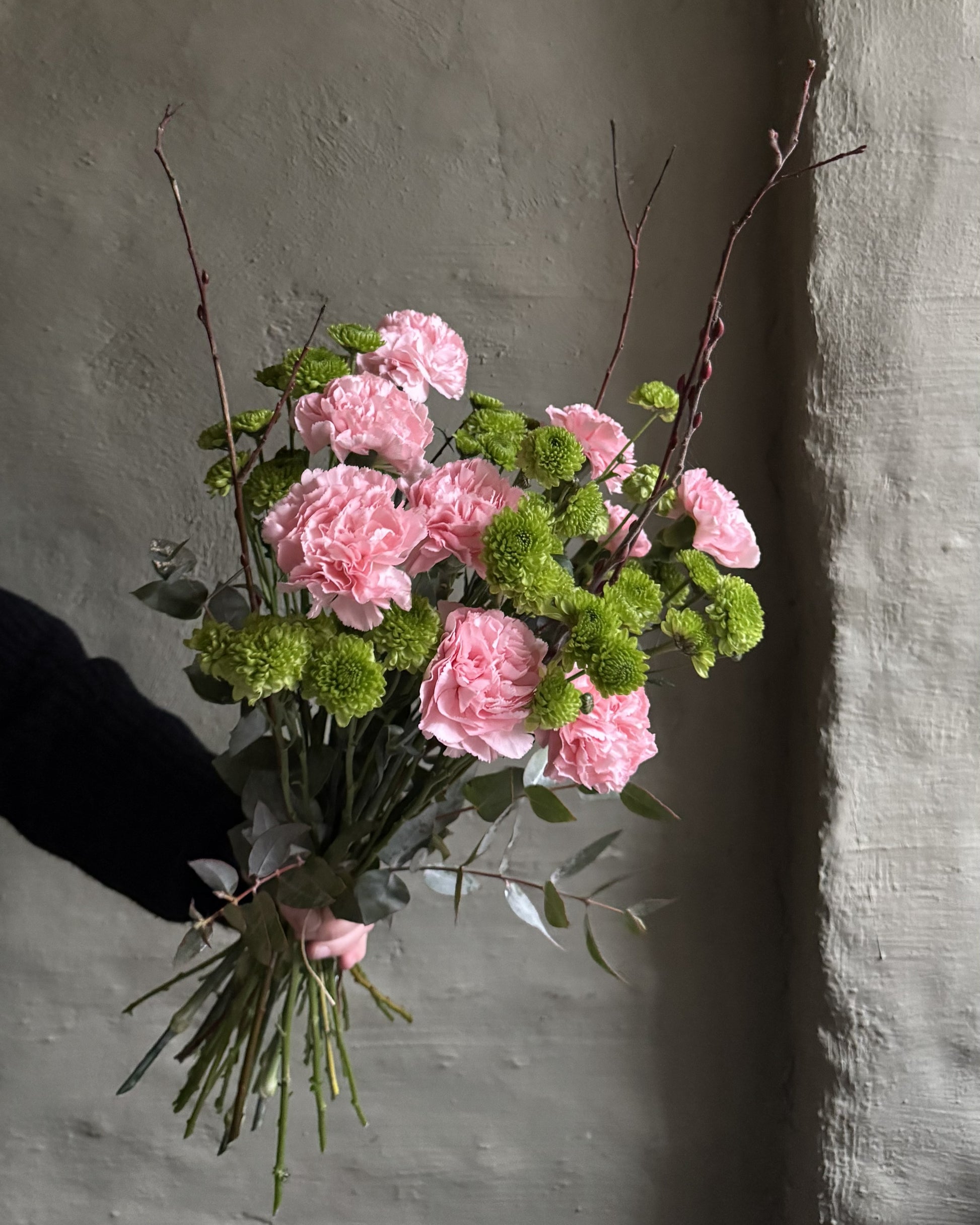 A bouquet of pink carnations and green pom-pom chrysanthemums with eucalyptus stems, held by a hand against a textured gray wall.