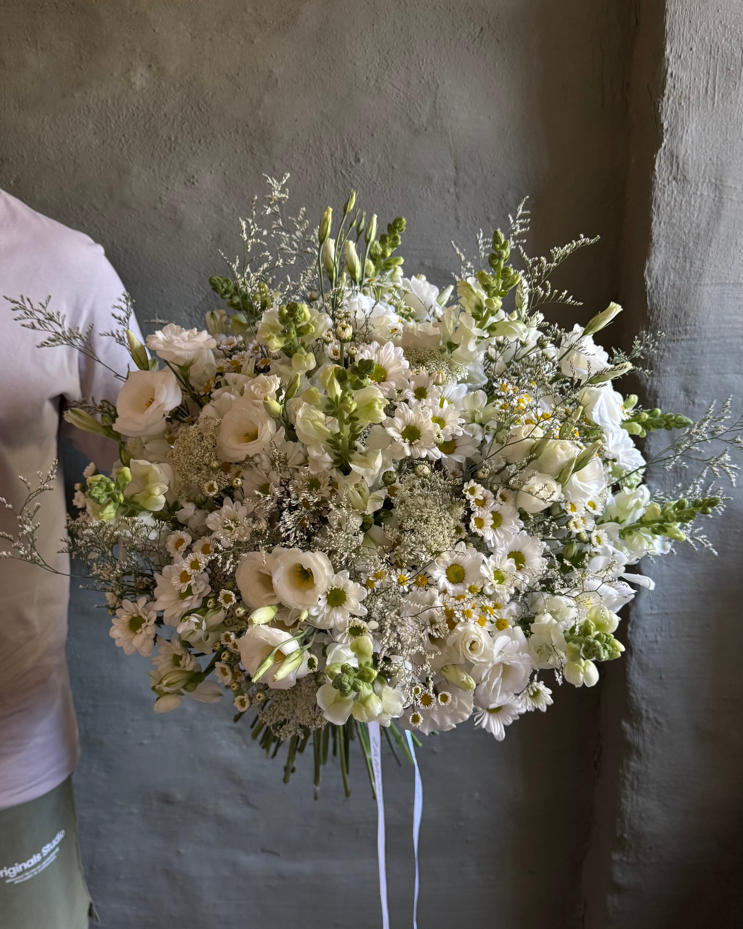 Person holding a large bouquet of white and green flowers against a plain wall.